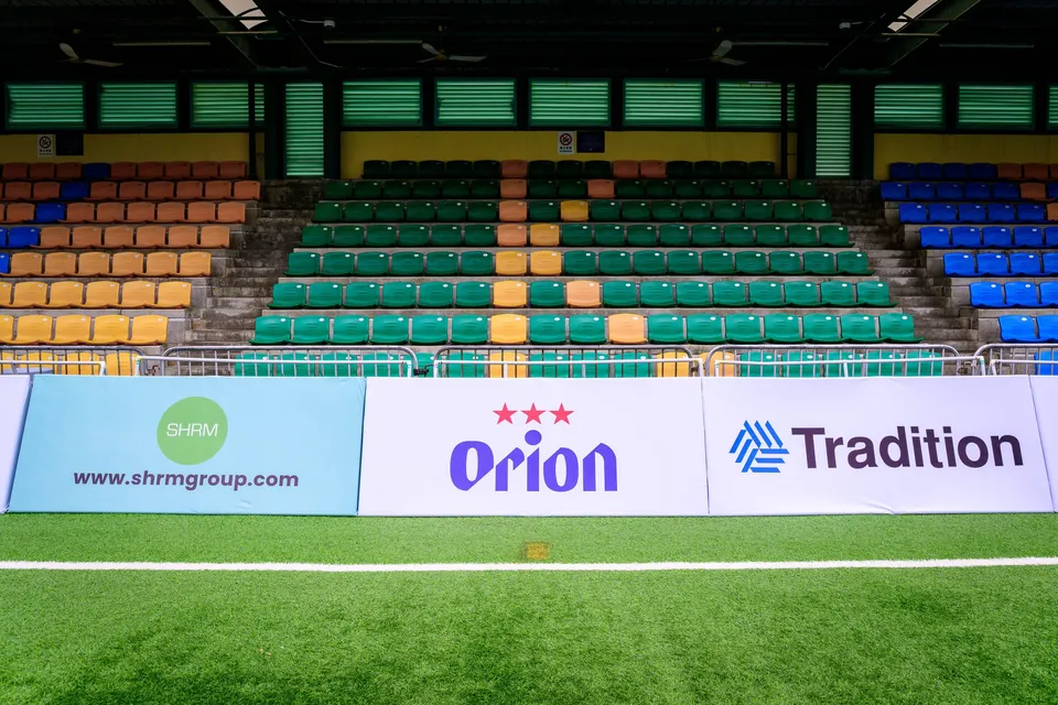 Empty stand with green and blue seats behind pitchside boards for SHRM, Orion and Tradition