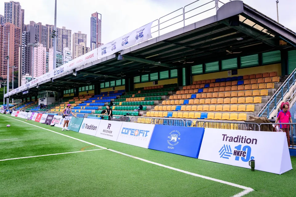 Covered grandstand along the touchline with sponsor boards and a Tradition HKFC 10s board, one spectator in pink