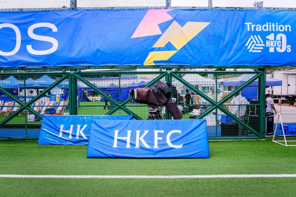 TV camera on the ground between HKFC boards, with a green fence and tents behind the pitch