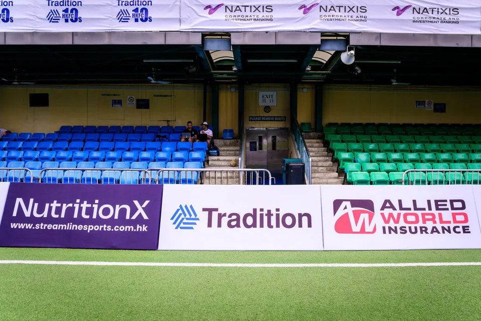 Empty stand with blue seating above NutritionX, Tradition and Allied World Insurance boards, two people seated