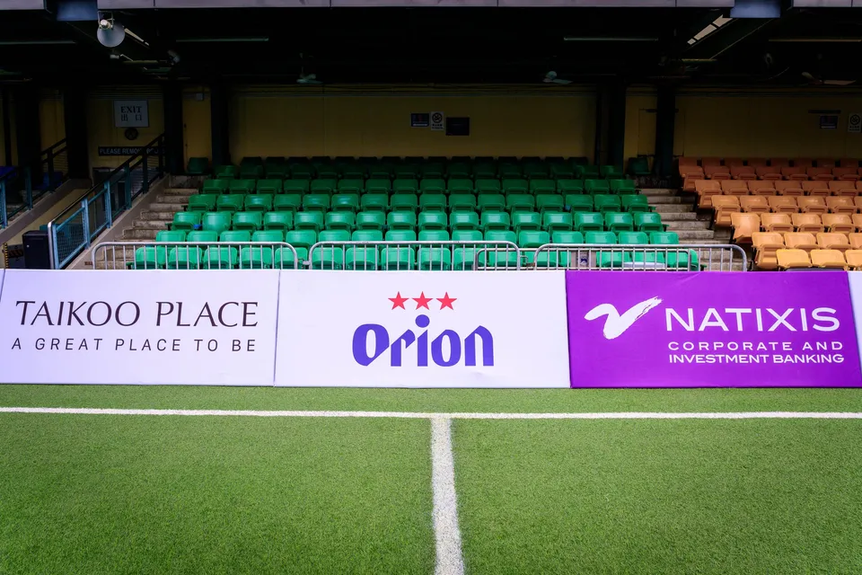 Pitchside boards for Taikoo Place, Orion and Natixis in front of green and orange stand seating
