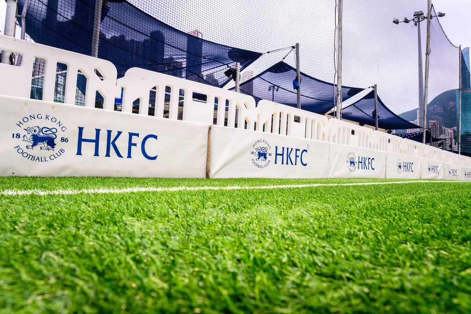 Low angle along the touchline of a row of sponsor boards, a Tradition HKFC 10s board and tall buildings beyond