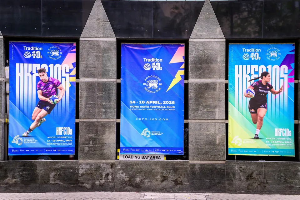 Lit poster frame on a stone wall showing three rugby players in action, with the Tradition HKFC 10s name and dates