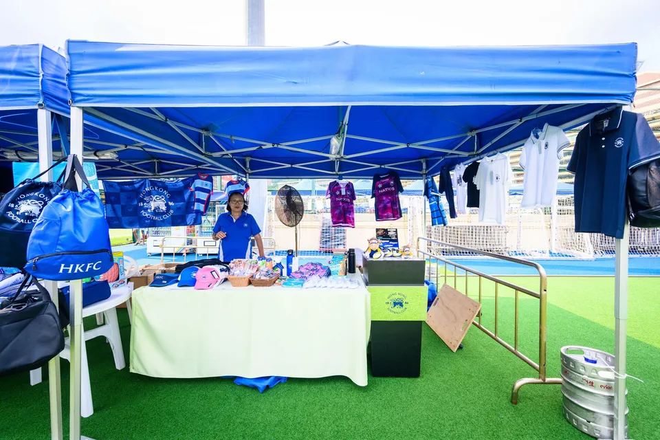 Merchandise stall under a blue canopy with club shirts, caps and bags on display and one helper at the table