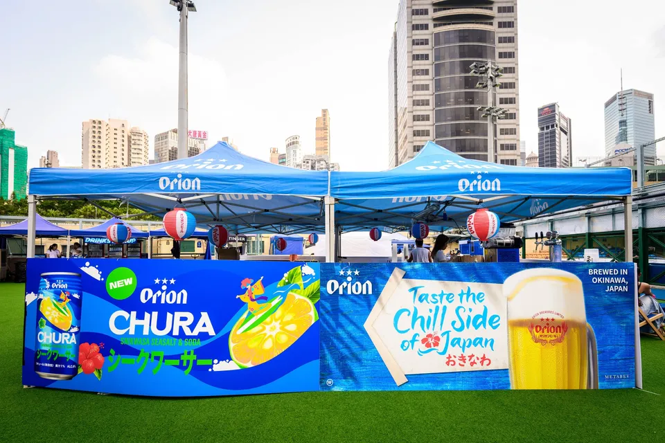 Orion beer stall with blue canopies and a Chura drink banner, hanging red lanterns on the pitch edge