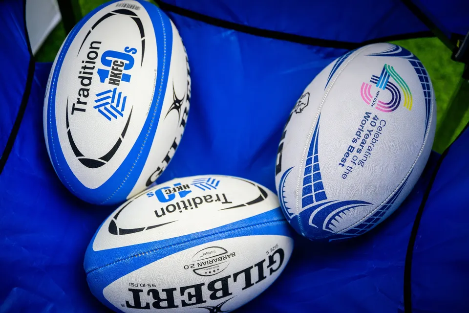 Three rugby balls on a blue cloth, two with Tradition HKFC 10s branding and one marked Gilbert Barbarians