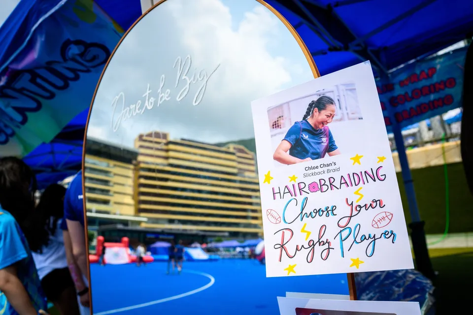 Hair braiding sign reading Choose Your Rugby Player, propped beside a mirror reflecting the pitch and stands