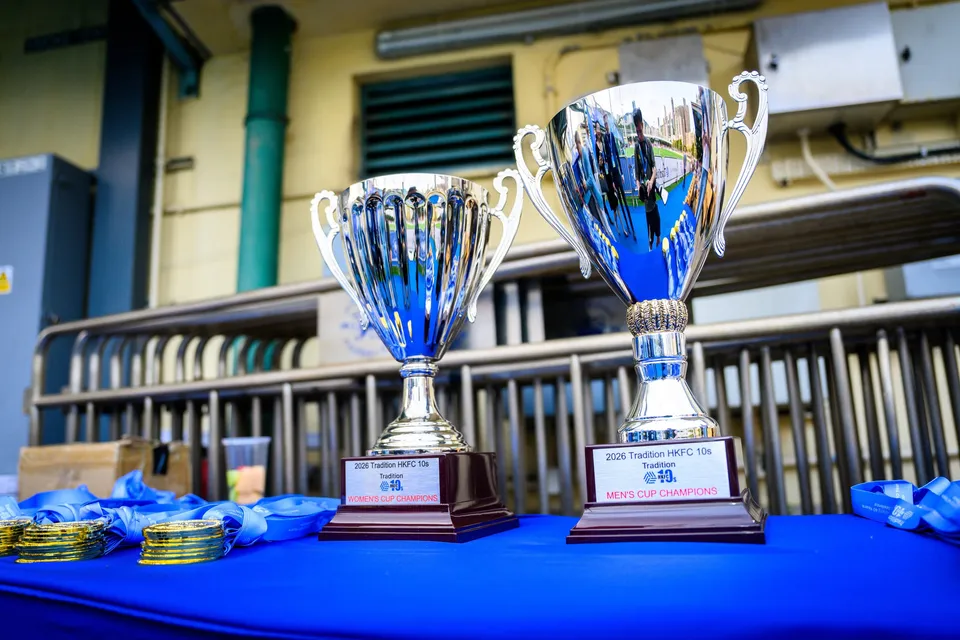 Two silver trophies on a blue table, one labelled Men's Cup Champions, with gold medals and ribbons beside them
